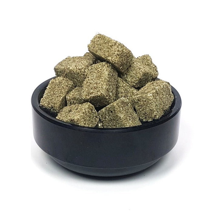 A pile of small, textured, olive-green herbal or supplement cubes resting in a small black bowl, set against a plain white background.