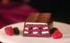 Slice of layered dessert on a plate with a box of Ulker Cikolatali Yas Pasta in the background. Ulker Milk Chocolate Cake with Velvety Red Velvet Base and Forest Berry Sauce – 13.9 oz (395 G)
