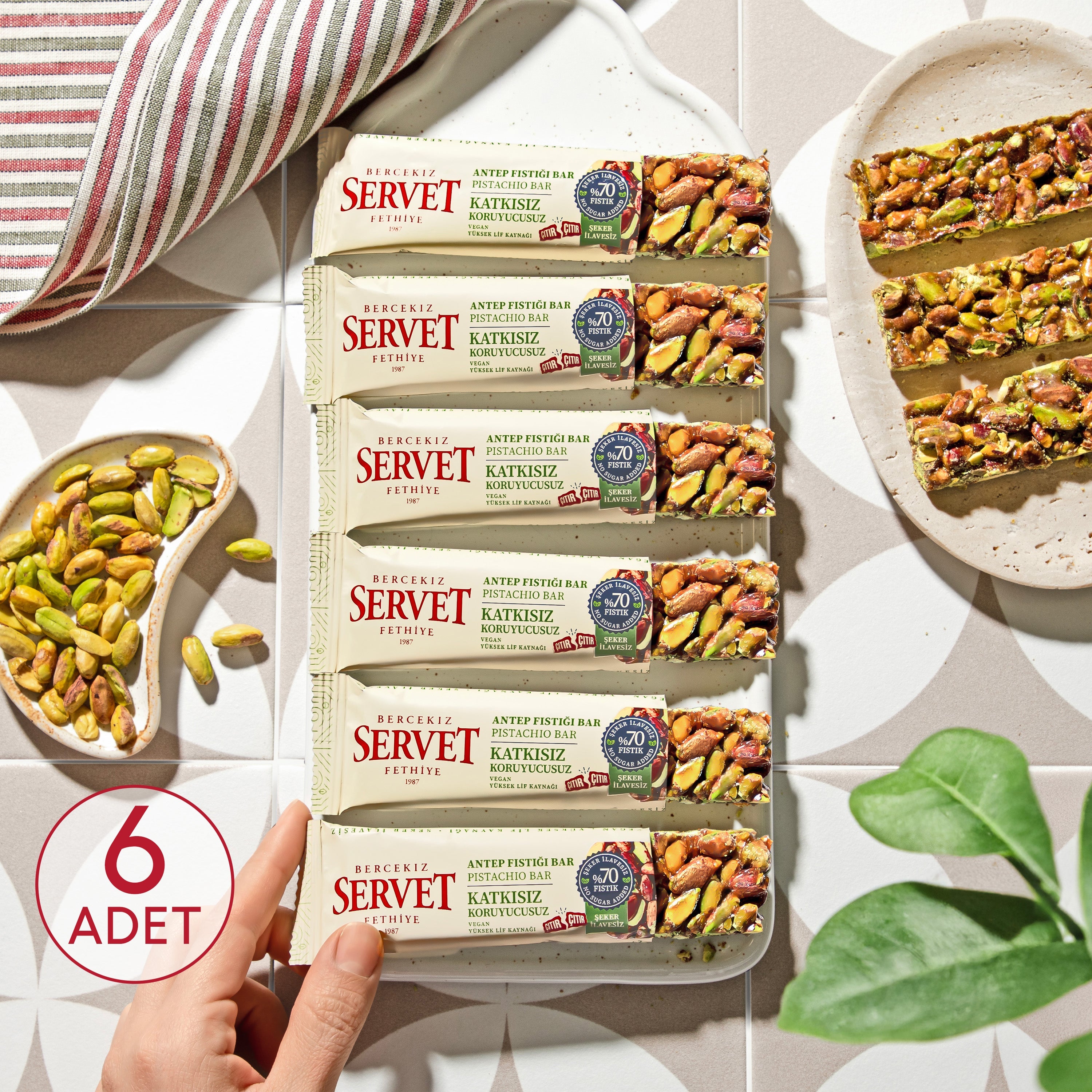 Packaging of Servet Antep Fındıklı Bar with a hand holding it, surrounded by pistachios and a plant.